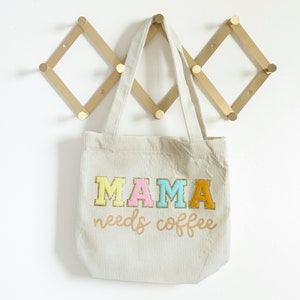 May include: A beige corduroy tote bag with the text "MAMA needs coffee" in colorful embroidered letters. The bag has two long straps and is hanging on a gold metal hook.