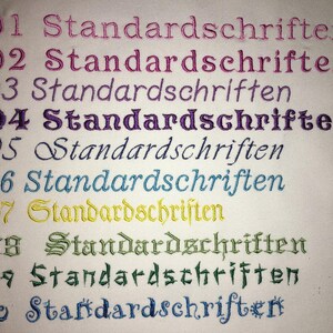 May include: A close-up of a white fabric with ten rows of embroidered text. Each row features the word "Standardschriften" in a different color, including pink, purple, teal, yellow, and green.