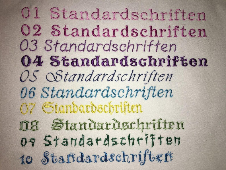 May include: A close-up of a white fabric with ten rows of embroidered text. Each row features the word "Standardschriften" in a different color, including pink, purple, teal, yellow, and green.