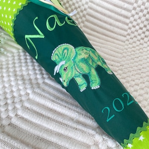 May include: A green and white school cone with a triceratops design. The cone is dark green with a green triceratops embroidered on the side. The word "Na" is embroidered in green cursive, and the year "202" is also embroidered.