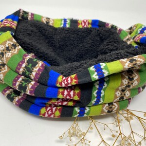 Op de afbeelding: Een kleurrijke, gepatroneerde nekwarmer met een zwarte fleece voering. De nekwarmer is gevouwen en ligt op een wit oppervlak.