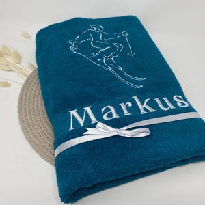 May include: Teal bath towel with white embroidered skier graphic and the name "Markus". A white ribbon with a bow is tied around the towel. The towel is folded and placed on a neutral surface.
