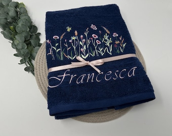 May include: A navy blue terry cloth towel featuring an embroidered design of colorful wildflowers, a small bee, and a butterfly. The name Francesca is embroidered in light pink script below the flowers, and a light pink ribbon is tied around it.