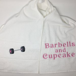 May include: White hooded towel with the text "Barbells and Cupcakes" embroidered in pink on the right side and a black and gray barbell embroidered on the left side.