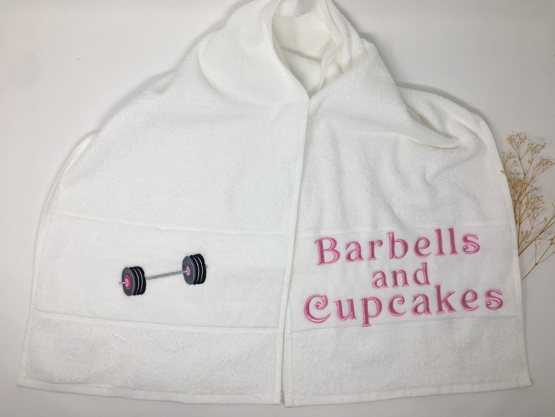 May include: White hooded towel with the text "Barbells and Cupcakes" embroidered in pink on the right side and a black and gray barbell embroidered on the left side.