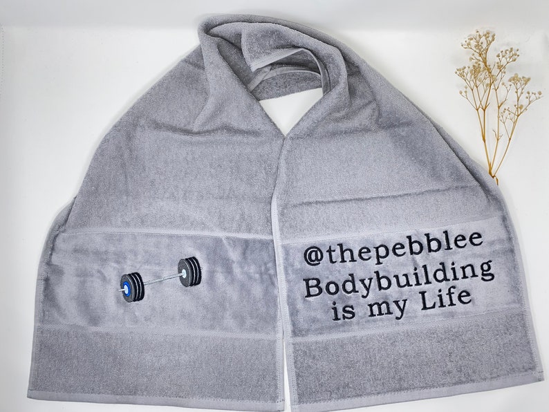 May include: Gray cotton towel with black embroidery. The embroidery includes a barbell with weights and the text "@thepebblee Bodybuilding is my Life".