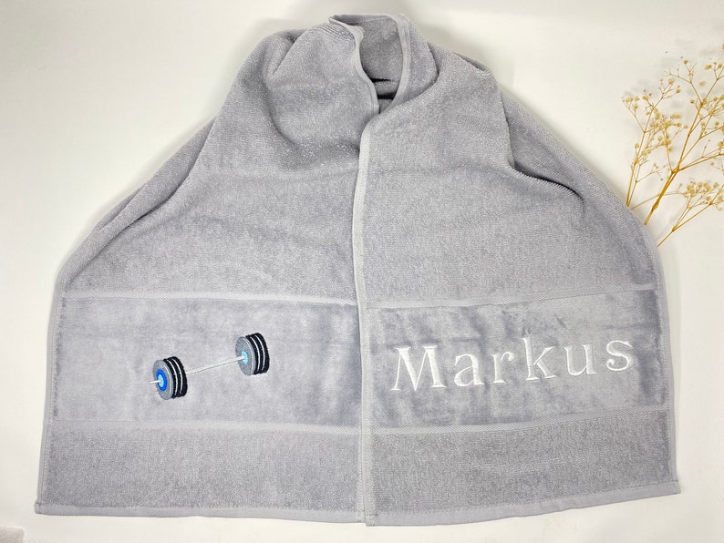 May include: Gray bath towel with a white barbell design and the name "Markus" embroidered on it.