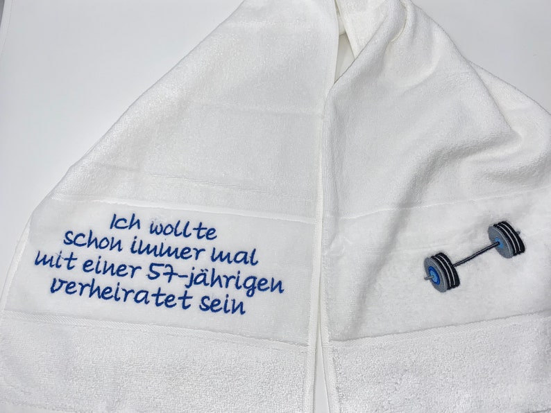 May include: White towel with blue embroidery that reads "Ich wollte schon immer mal mit einer 57-j&auml;hrigen verheiratet sein" and a gray barbell.