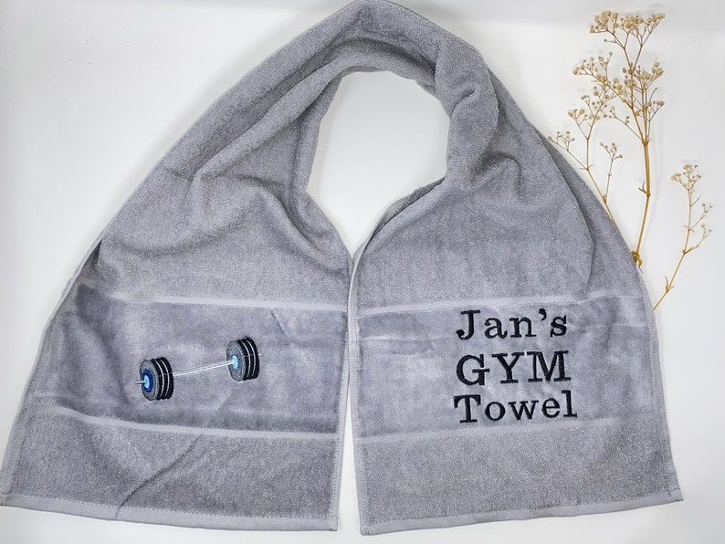 May include: Gray gym towel with the text "Jan's GYM Towel" and a black and white embroidered image of a barbell.