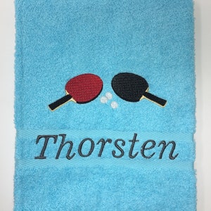 May include: A light blue towel with a black and red embroidered table tennis paddle design and the name "Thorsten" embroidered below.