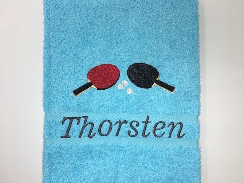 May include: A light blue towel with a black and red embroidered table tennis paddle design and the name "Thorsten" embroidered below.