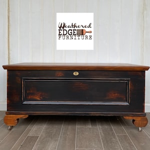 May include: A large, antique wooden chest with a black painted front and a brown stained top. The chest has a keyhole and sits on small wheels. The text "Weathered EDGE FURNITURE" is displayed on a white background above the chest.