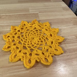 Könnte beinhalten: Eine gelbe gehäkelte Tischdecke mit einem runden Design. Die Tischdecke ist aus Garn gefertigt und hat eine zarte, spitzenartige Optik.