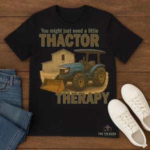 Op de afbeelding: Zwart t-shirt met een afbeelding van een blauwe tractor en de tekst "You might just need a little THACTOR THERAPY". Het ontwerp bevat een schuur en een tractor met een voorlader.