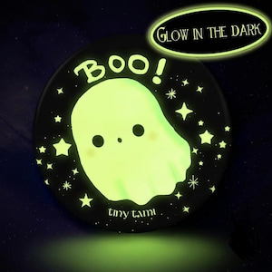 Könnte beinhalten: Ein runder schwarzer Anstecker mit einem leuchtend hellgrünen Geist in der Mitte, darüber der Text BOO! und darunter tiny tami. Das Design enthält verschiedene Sternformen um den Geist herum und ein Etikett mit GLOW IN THE DARK.