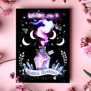 Könnte beinhalten: Schwarze Geburtstagskarte mit magischem Thema. Sie zeigt eine Zaubertrankflasche mit rosa und violettem Dampf, Kristallen, Halbmonden und dem Text "Wishing you a Magical Birthday."