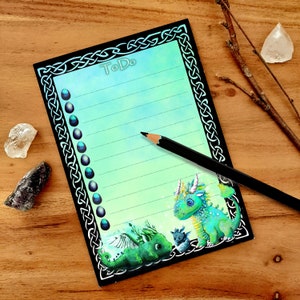 Può includere: Un blocco note nero e verde per la lista delle cose da fare con un bordo a nodo celtico. Il blocco note presenta un'illustrazione di drago verde e blu con il testo "To Do" in alto.