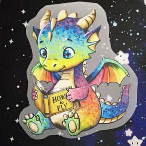 Könnte beinhalten: Ein gestanzter Aufkleber mit einem bunten Cartoon-Baby-Drachen mit blauen, grünen und gelben Schuppen. Der Drache hat kleine Flügel, Hörner und einen Schwanz und hält ein gelbes Buch mit dem Text "HOW to FLY" auf dem vorderen Einband.