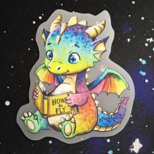 Könnte beinhalten: Ein gestanzter Aufkleber, der einen bunten, regenbogenfarbenen Drachen mit großen blauen Augen zeigt. Der Drache sitzt und hält ein gelbes Buch mit dem aufgedruckten Text "HOW to FLY" auf dem Einband. Der Drache hat kleine Hörner und Flügel.
