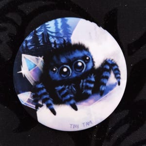Op de afbeelding: Een ronde button met een illustratie van een blauwe springspin. De achtergrond toont een bos en kristallen. De tekst "TINY TAM" staat onderaan. De spin heeft gestreepte poten en een harig lichaam.