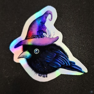 Op de afbeelding: Een holografische sticker met een zwarte kraai die een paarse heksenhoed draagt. De hoed is versierd met een kleine spin, kristallen en een regenboogkleurige rand. De kraai heeft een blauw oog en blauwe schaduwen op zijn veren.
