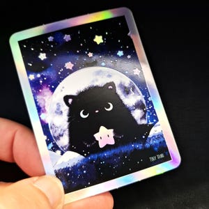 Könnte beinhalten: Ein holografischer Aufkleber mit einer schwarzen Katze, die einen Stern vor einem Vollmond hält. Der Hintergrund ist ein Sternenhimmel mit Pastellfarben. Der Aufkleber hat eine rechteckige Form mit abgerundeten Ecken und dem Text "TINY TAMI".
