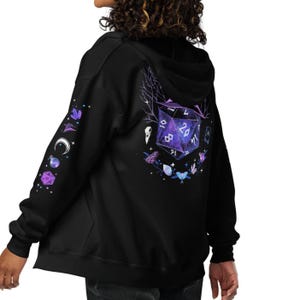 Può includere: Felpa con cappuccio nera con zip e design a tema fantasy. La parte posteriore presenta un grande dado viola a venti facce con altri elementi magici. La manica sinistra ha disegni celesti e geometrici aggiuntivi. La felpa è realizzata in materiale morbido.