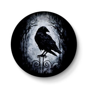 Könnte beinhalten: Runde, schwarze Grafik mit einem schwarzen Raben, der auf einem dekorativen Tor sitzt. Der Hintergrund zeigt einen nebligen Wald mit kahlen Bäumen. Der Text "TINY TAMI" befindet sich am unteren Rand des Tors.
