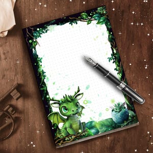 Op de afbeelding: Een fantasie-thema notitieboek met een groene drakenillustratie en een zwarte vulpen. Het notitieboek heeft een gestippeld rasterpatroon en een decoratieve rand van groene bladeren en wijnranken. De draak is afgebeeld met grote ogen en vleugels.