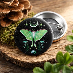 Könnte beinhalten: Ein runder Ansteckbutton mit schwarzem Hintergrund und einer zentralen Illustration eines grünen Mondspinners. Das Design zeigt eine Mondsichel, kleine weiße Sterne, zwei Pilze, grünes Blattwerk und eine Ansammlung von Kristallen am unteren Rand.
