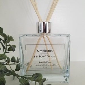May include: A square clear glass reed diffuser with a silver cap and wooden reeds. The label reads Simplicities, Bamboo & Coconut, Reed Diffuser, handcrafted in Chardon, Ohio, 7.5 oz. The bottle is positioned with light-colored wooden reeds inserted.