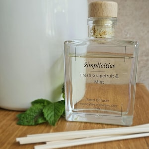 May include: A clear glass reed diffuser bottle with a light-colored wooden stopper. The label reads Simplicities Fresh Grapefruit & Mint Reed Diffuser handcrafted in Chardon, Ohio 5 oz. Several white reed sticks are placed in front of the bottle.