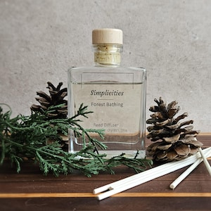 May include: A clear glass reed diffuser bottle with a wooden cap, labeled "Simplicities Forest Bathing." The bottle is surrounded by pine cones, greenery, and white diffuser sticks, all arranged on a wooden surface.
