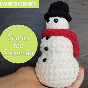May include: A crocheted snowman named Charles wearing a black hat and a red scarf. The snowman is sitting in a person's hand. The text "PDF Crochet Pattern Charles the Snowman" is on a green circle.