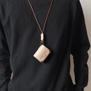 May include: A light beige leather wallet with a matching lanyard. The wallet is rectangular and appears to be closed. The lanyard is a thin, light brown leather cord. The text "TANGENT LINEAR" is visible on the wallet.