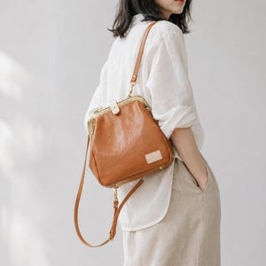 May include: A brown leather crossbody bag with a kiss-lock closure and a long adjustable strap. The bag has a rectangular shape with rounded corners and a small rectangular patch on the front. The hardware is gold-toned.