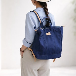 May include: A navy blue canvas tote bag with tan leather straps and accents. The bag is worn as a backpack, featuring a rectangular shape with a top handle and a small leather patch. The bag is carried by a person wearing a light blue shirt and tan pants.