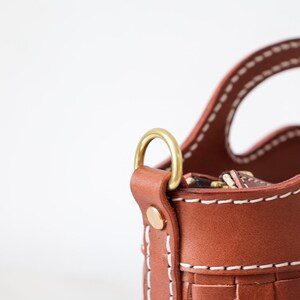 Puede incluir: Primer plano de un bolso de mano de cuero marr&oacute;n con un dise&ntilde;o tejido. El bolso presenta costuras blancas, un anillo de metal dorado y un asa. El bolso es de color marr&oacute;n claro.