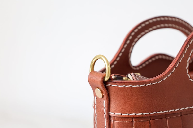 Puede incluir: Primer plano de un bolso de mano de cuero marr&oacute;n con un dise&ntilde;o tejido. El bolso presenta costuras blancas, un anillo de metal dorado y un asa. El bolso es de color marr&oacute;n claro.