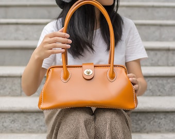 May include: A brown leather handbag with a top flap closure and a single strap. The bag has a stitched design and a gold metal clasp.