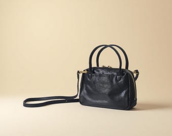 May include: A black leather handbag with a top handle and a detachable shoulder strap. The bag has a rounded shape with a zipper closure and gold-toned hardware. The bag is set against a neutral background.