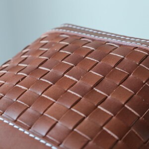 Puede incluir: Primer plano de un bolso de cuero marr&oacute;n con un patr&oacute;n tejido. El bolso presenta un dise&ntilde;o de tejido de cesta con costuras blancas a lo largo de los bordes. El cuero tiene un tono rico y c&aacute;lido.