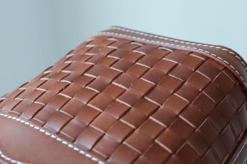 Puede incluir: Primer plano de un bolso de cuero marr&oacute;n con un patr&oacute;n tejido. El bolso presenta un dise&ntilde;o de tejido de cesta con costuras blancas a lo largo de los bordes. El cuero tiene un tono rico y c&aacute;lido.