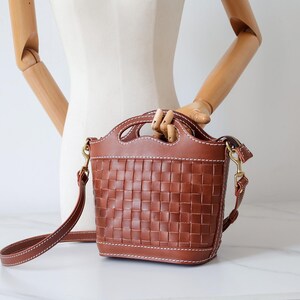 Puede incluir: Bolso de mano de cuero marr&oacute;n con dise&ntilde;o de cesta tejida. El bolso tiene un asa superior y una correa para el hombro desmontable. El bolso mide aproximadamente 20 cm de alto y 18 cm de ancho.