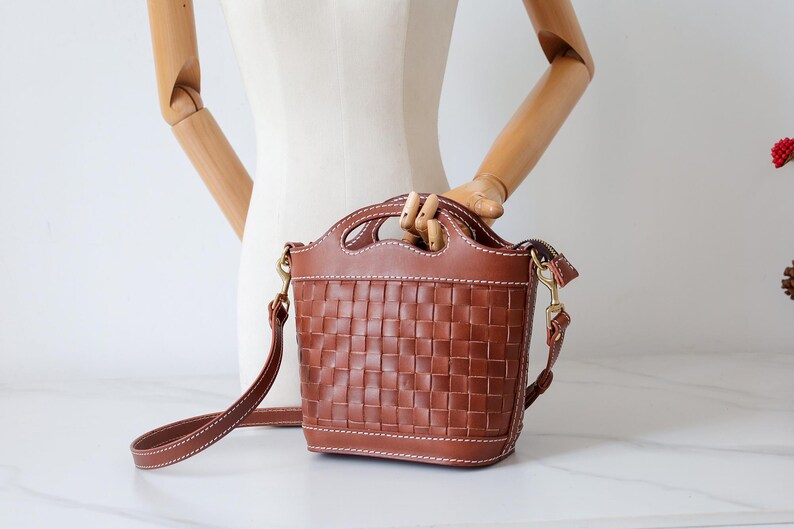 Puede incluir: Bolso de mano de cuero marr&oacute;n con dise&ntilde;o de cesta tejida. El bolso tiene un asa superior y una correa para el hombro desmontable. El bolso mide aproximadamente 20 cm de alto y 18 cm de ancho.
