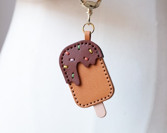 May include: A brown leather ice cream keychain with a chocolate topping and colourful sprinkles. The keychain has a gold metal ring and a brown leather stick.