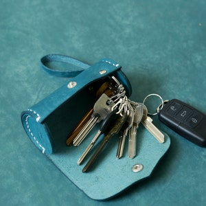 May include: A teal blue leather key holder with a zip closure and a silver metal snap. The key holder is open and filled with keys, including a car key fob.