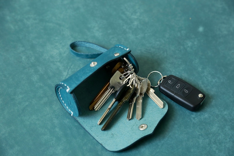 May include: A teal blue leather key holder with a zip closure and a silver metal snap. The key holder is open and filled with keys, including a car key fob.