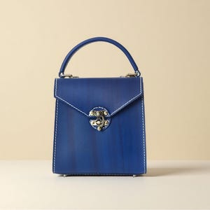 May include: A square, deep blue handbag with a top handle and a silver clasp. The bag has white stitching and a flap closure. The bag is on a white surface against a neutral background.