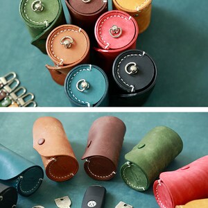 May include: An assortment of leather key holders in various colours, including brown, green, blue, red, and yellow. The key holders are rolled up and have a button closure. There are also several keys and a car key fob on a blue surface.